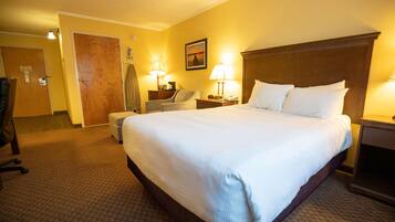 A hotel room with a large bed, a desk, a chair, a lamp, and a door.