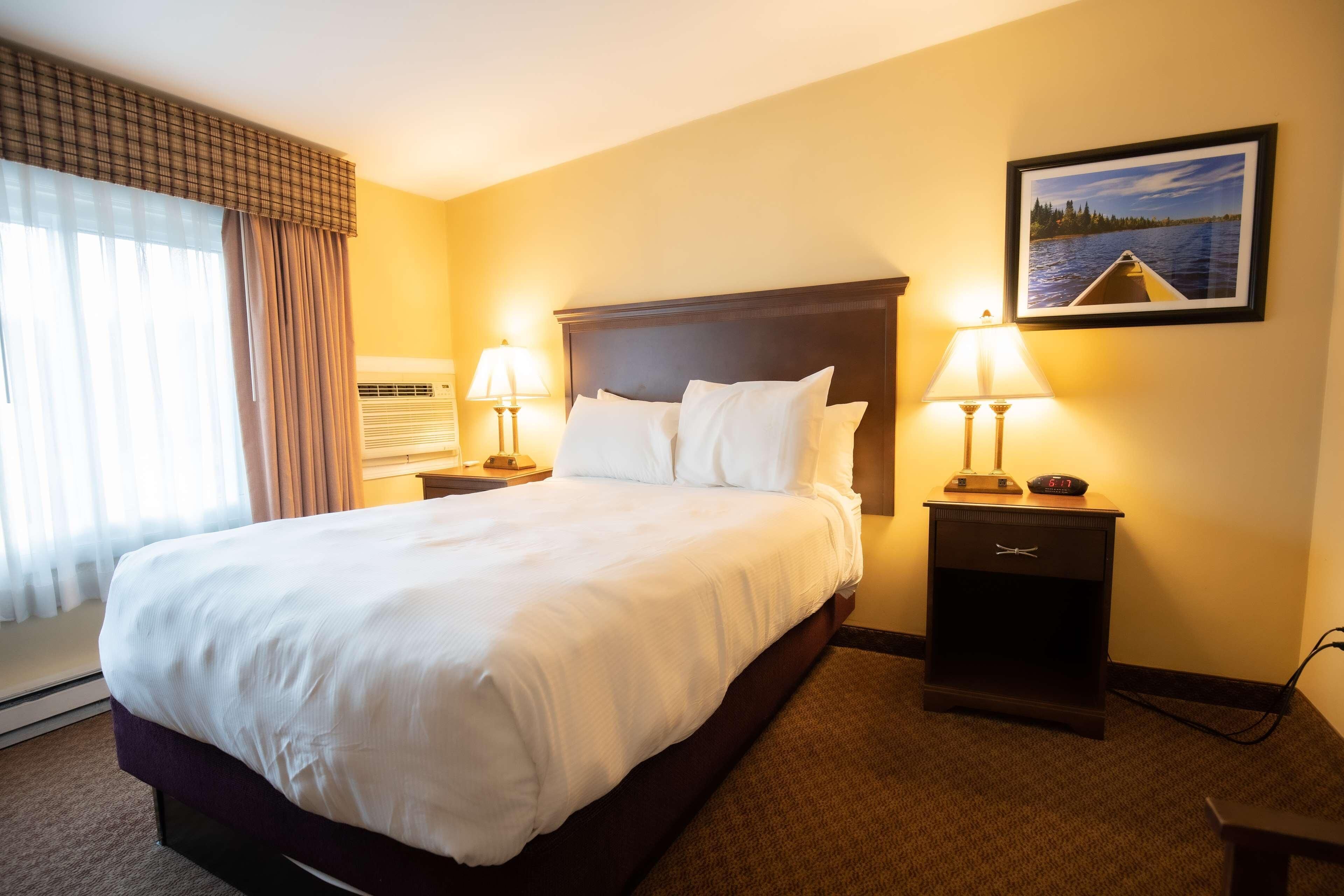 A hotel room with a large bed, two bedside tables with lamps, a wall-mounted clock, and a framed picture.