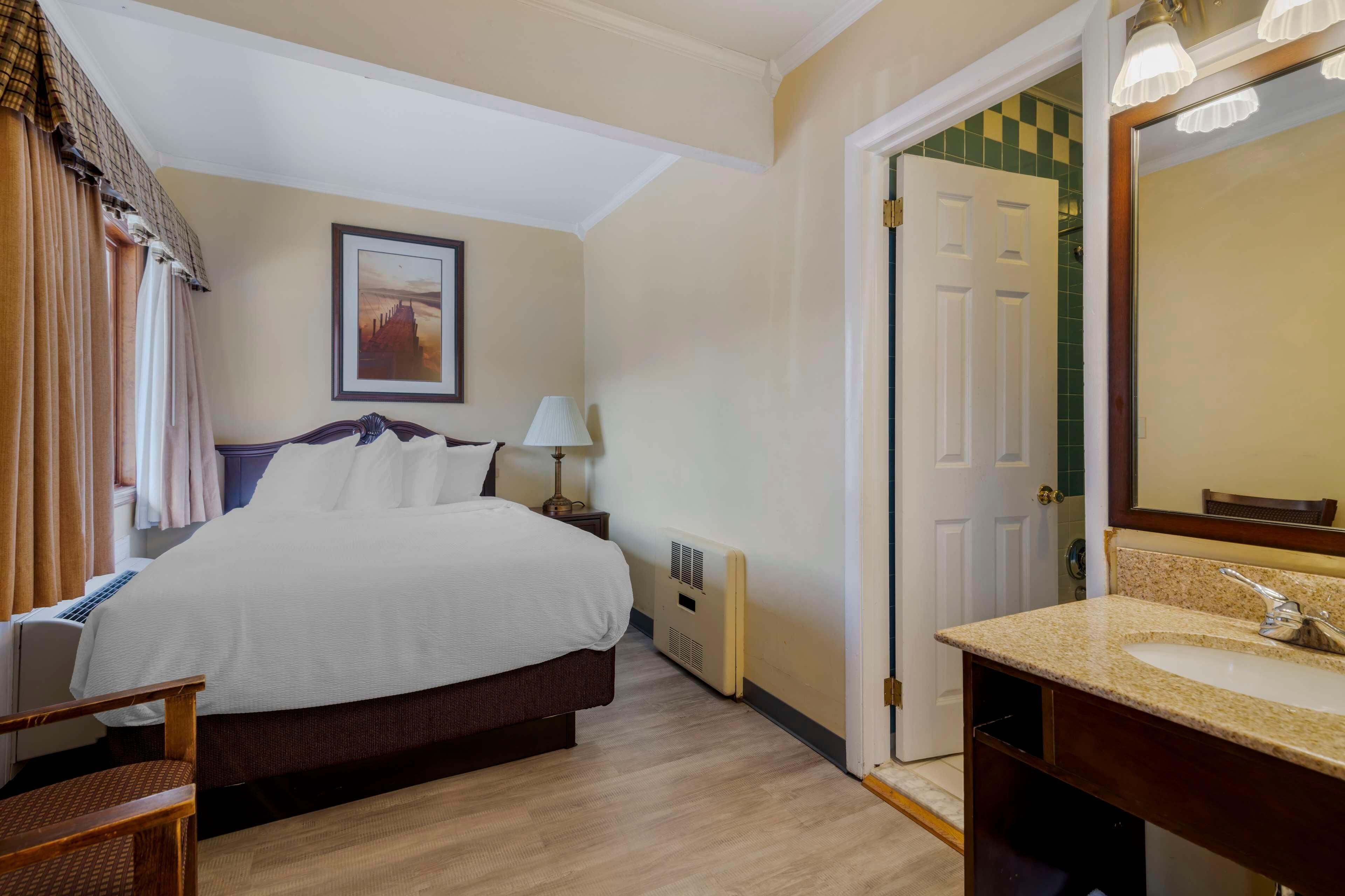 A hotel room with a bed, a bedside table with a lamp, a bathroom with a sink and mirror, and a door leading to another room.
