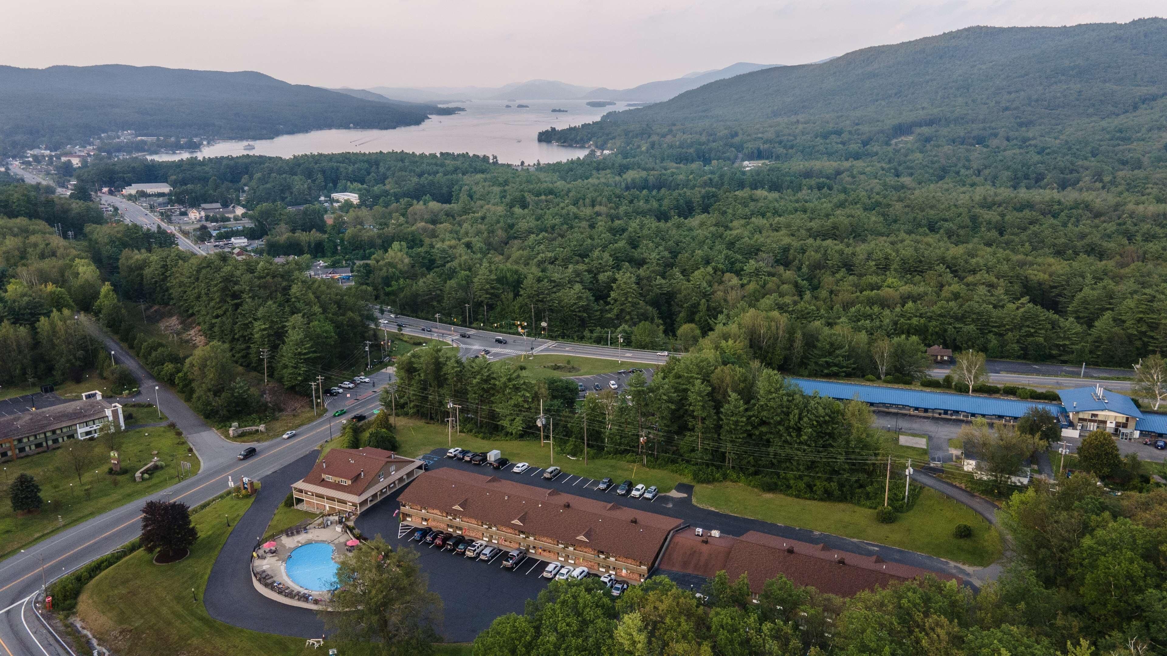 A hotel with a swimming pool, parking lot, and a view of a lake and forested hills.
