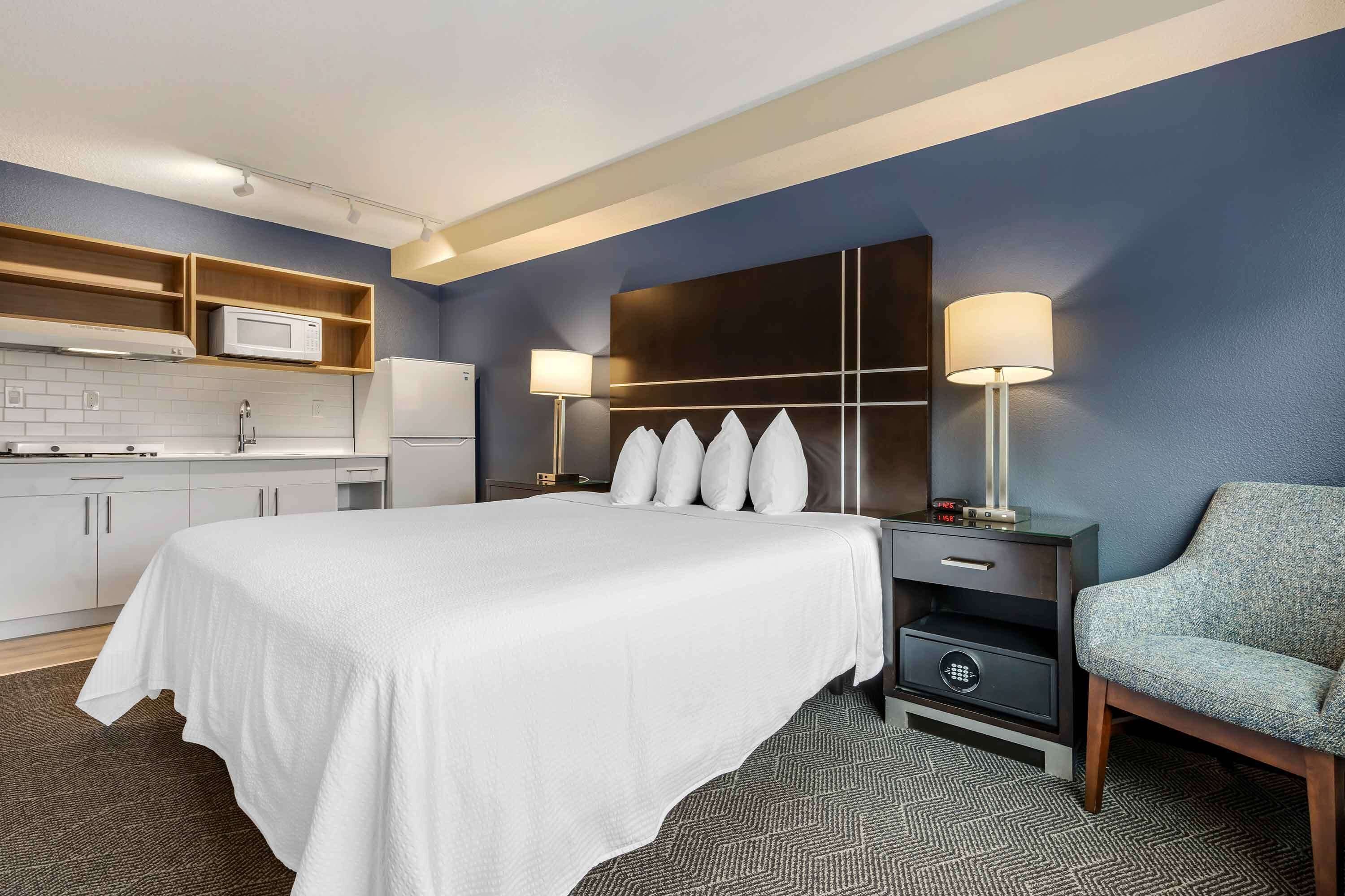 A hotel room with a large bed, a desk with a lamp, a chair, a microwave, and a small kitchenette.