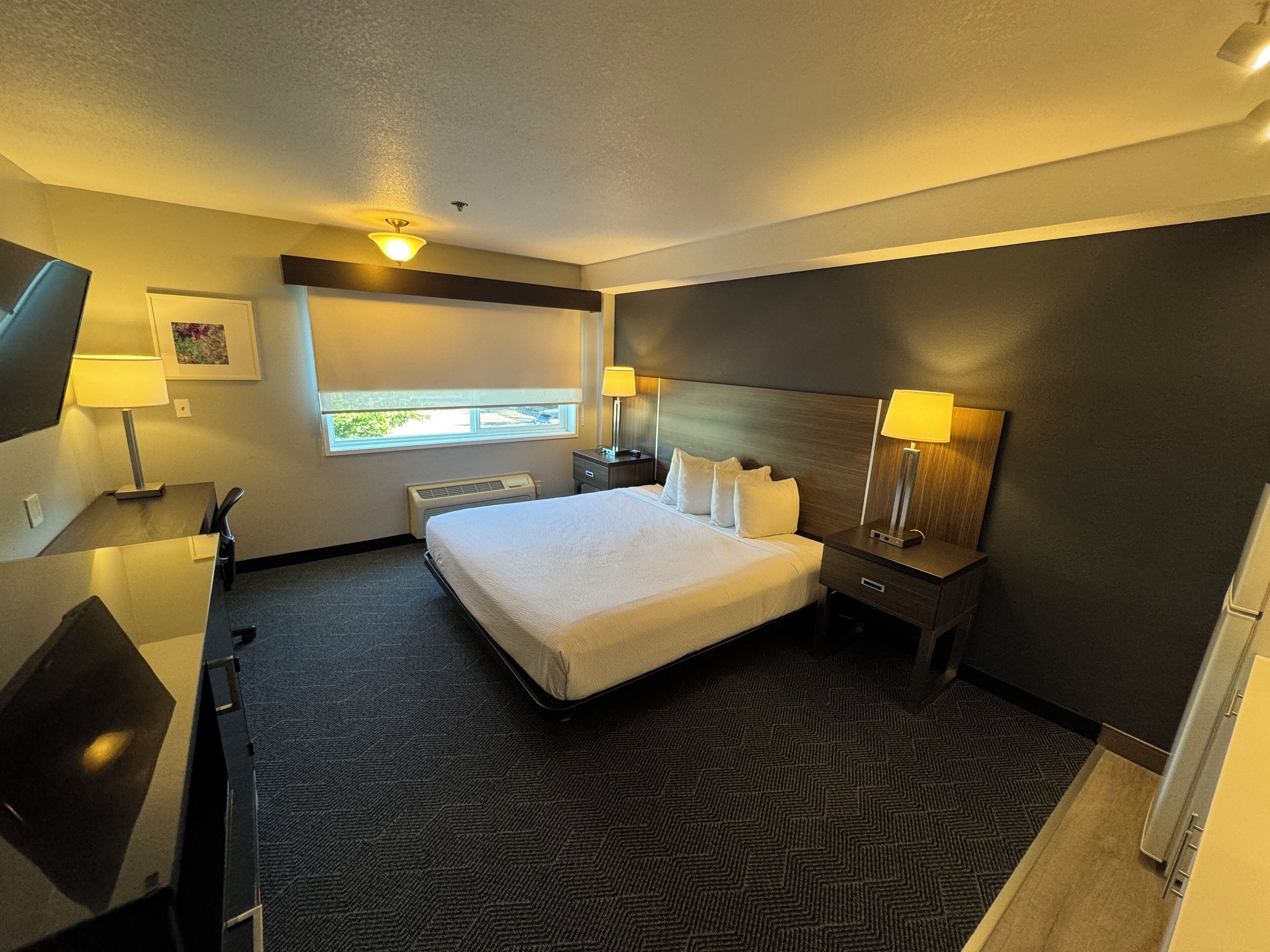 Une chambre d’hôtel avec un grand lit, des tables de chevet avec des lampes, un bureau, une chaise, une fenêtre donnant sur un beau paysage et un luminaire au plafond.