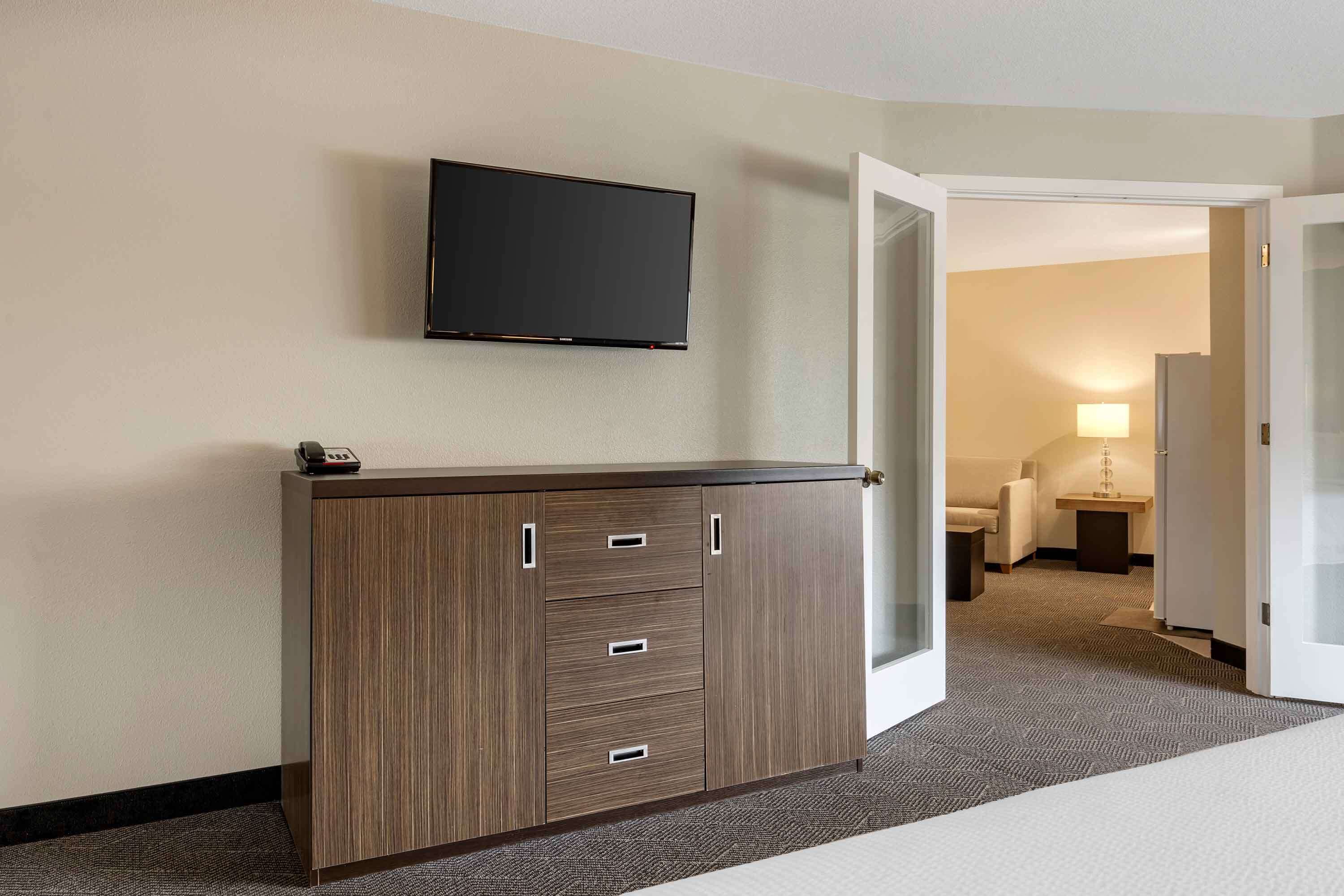 Habitación de hotel con televisor de pantalla plana montado en la pared, un aparador de madera con cajones y una puerta abierta que da a otra habitación con una cama y una mesita de noche.