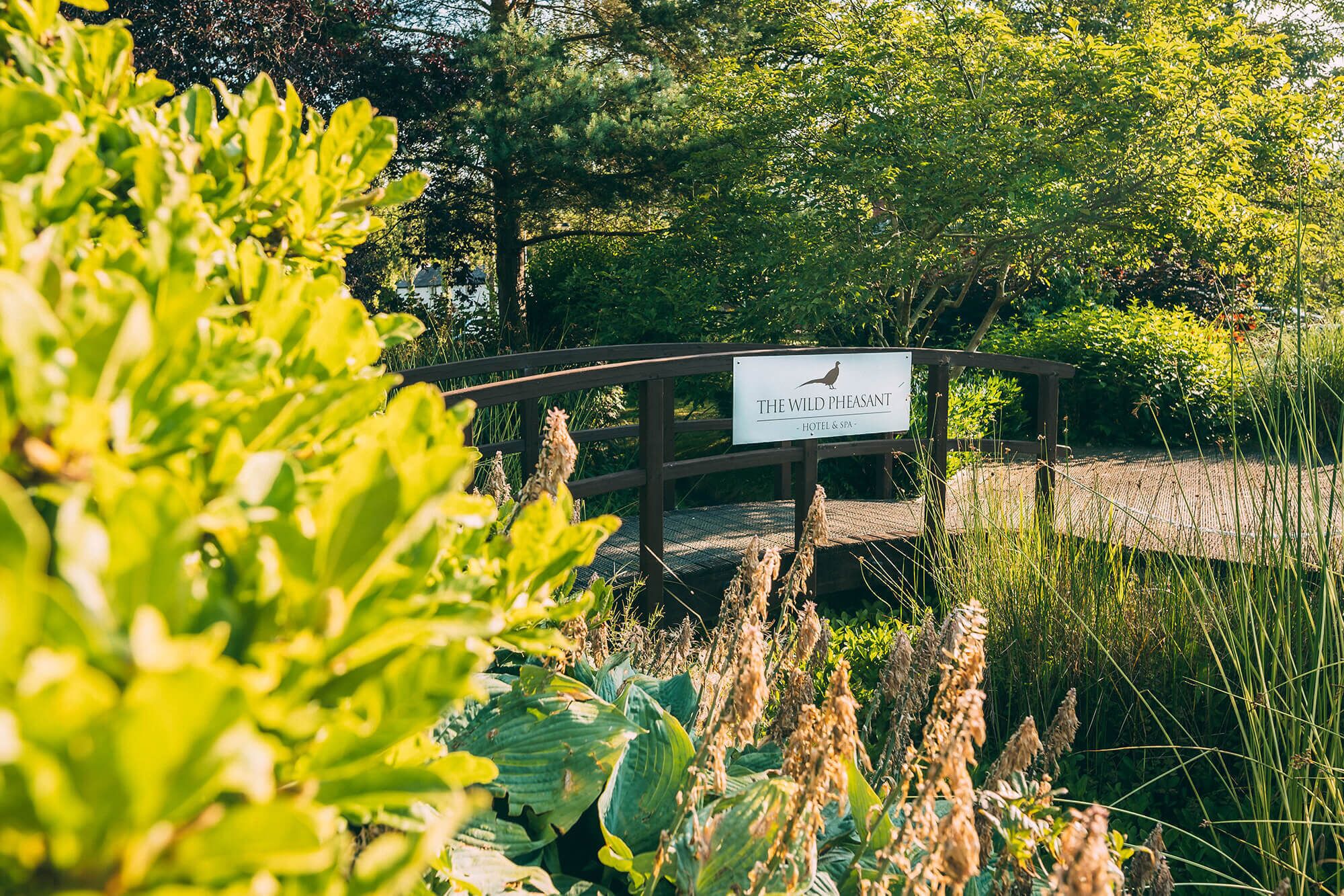 A wooden bridge with a sign for "The Wild Pheasant Hotel & Spa" in a lush garden setting.