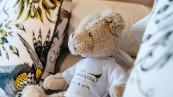 Een teddybeer met een shirt met het logo van 'The Pheasant Hotel & Spa', zittend op een bed met gestreepte kussens.