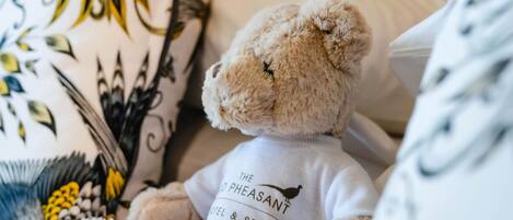 A teddy bear wearing a shirt with 'The Pheasant Hotel & Spa' logo, sitting on a bed with patterned pillows.