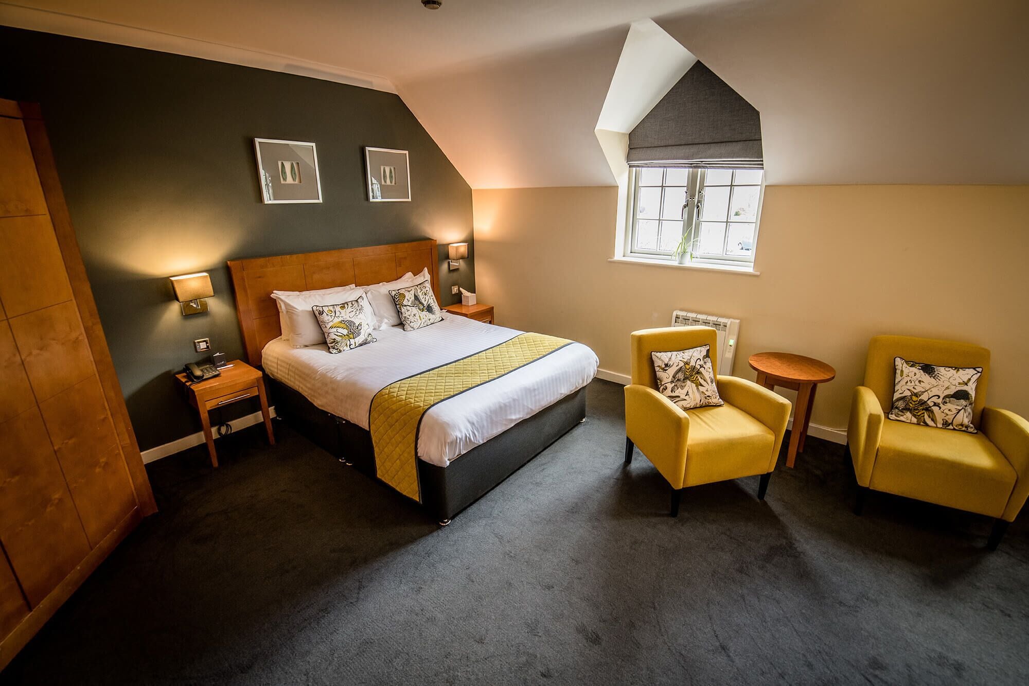 A bedroom with a large bed, two bedside tables, a small window, and two yellow armchairs.