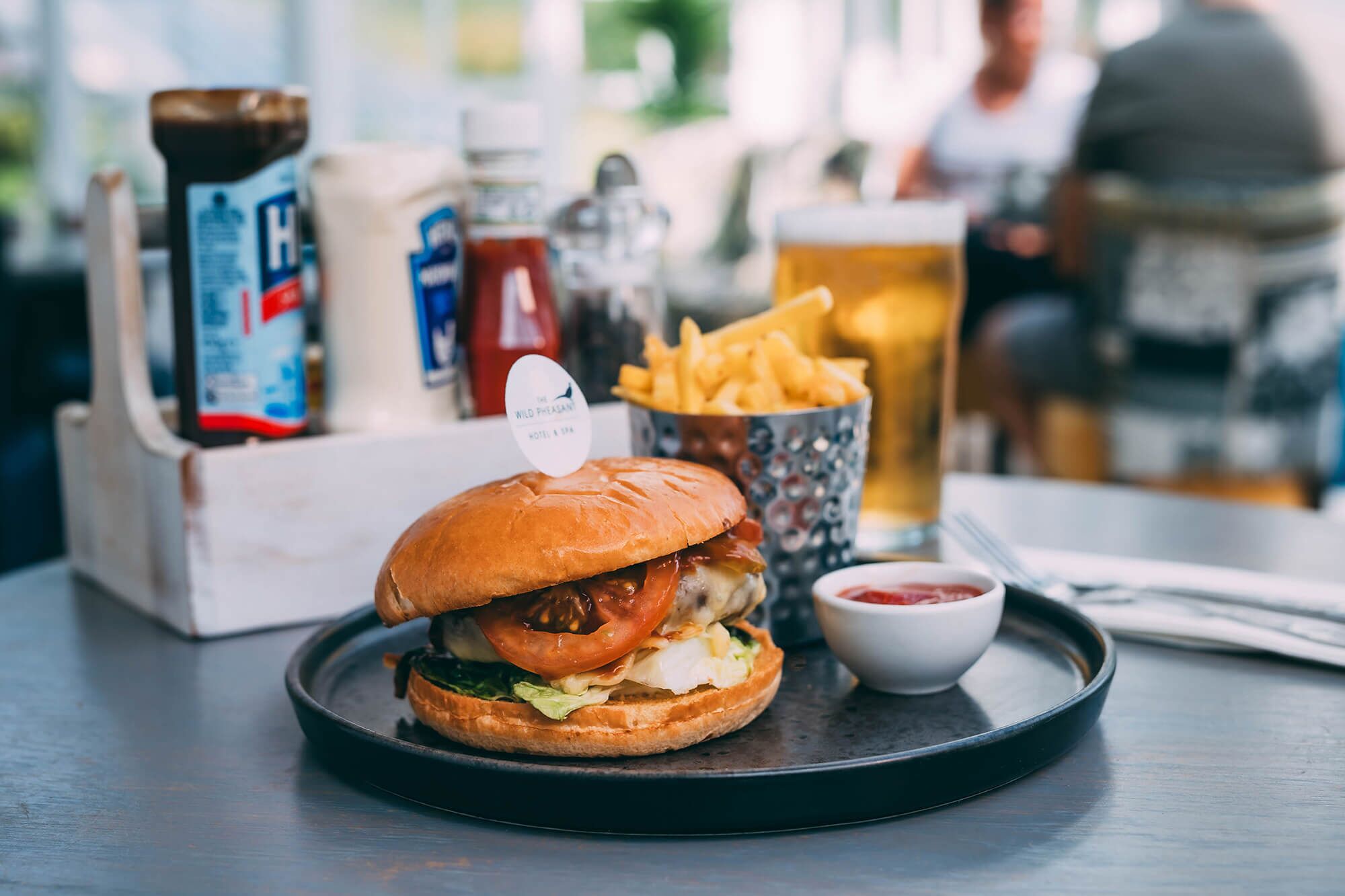 Una hamburguesa con papas fritas y kétchup sobre una bandeja, un vaso de cerveza y salsas en una mesa.