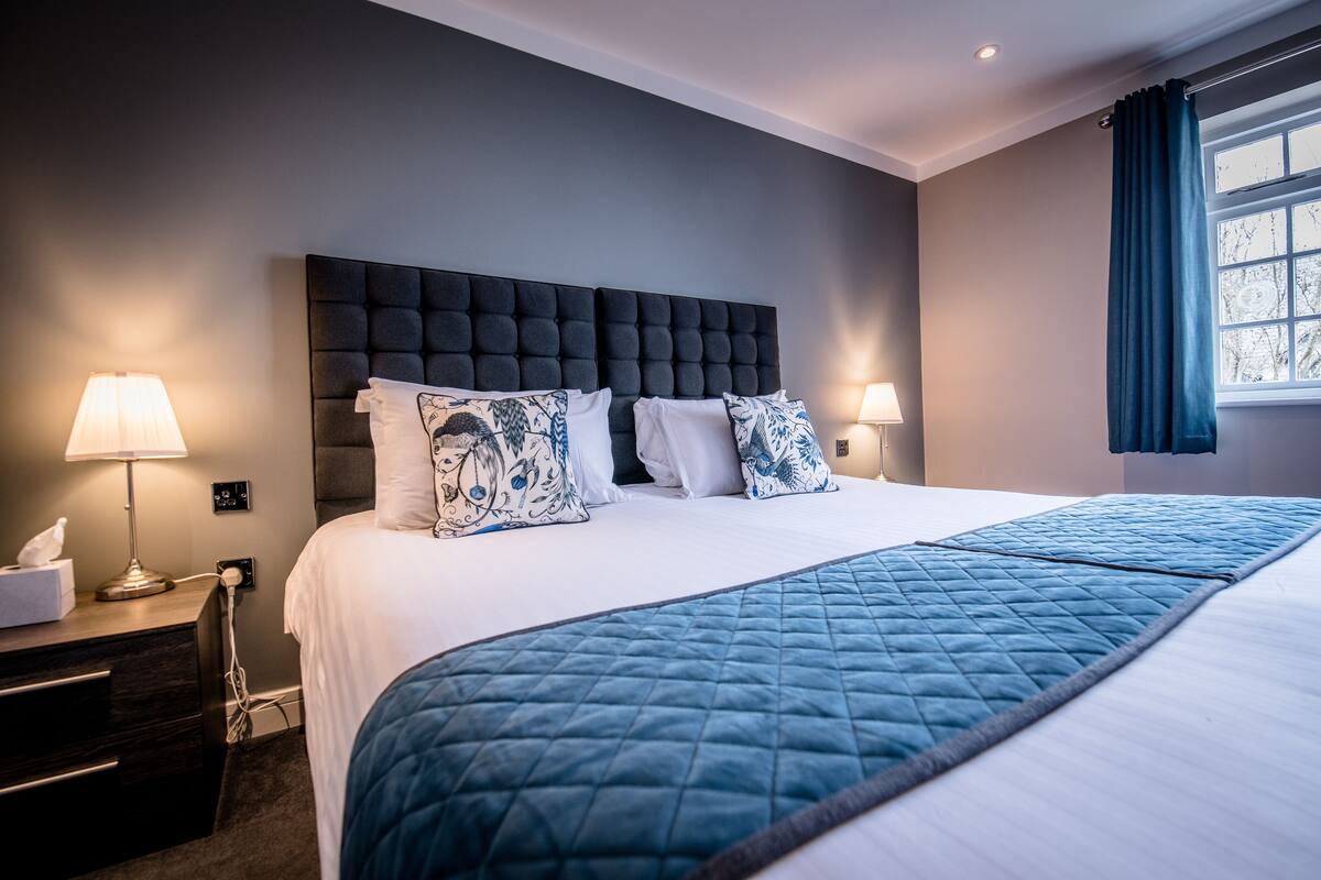 Una habitación de hotel con una cama grande, dos mesitas de noche con lámparas, un cabecero oscuro y una ventana con cortinas.