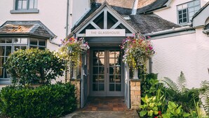 A traditional glasshouse entrance with a sign, surrounded by lush greenery and a brick pathway.