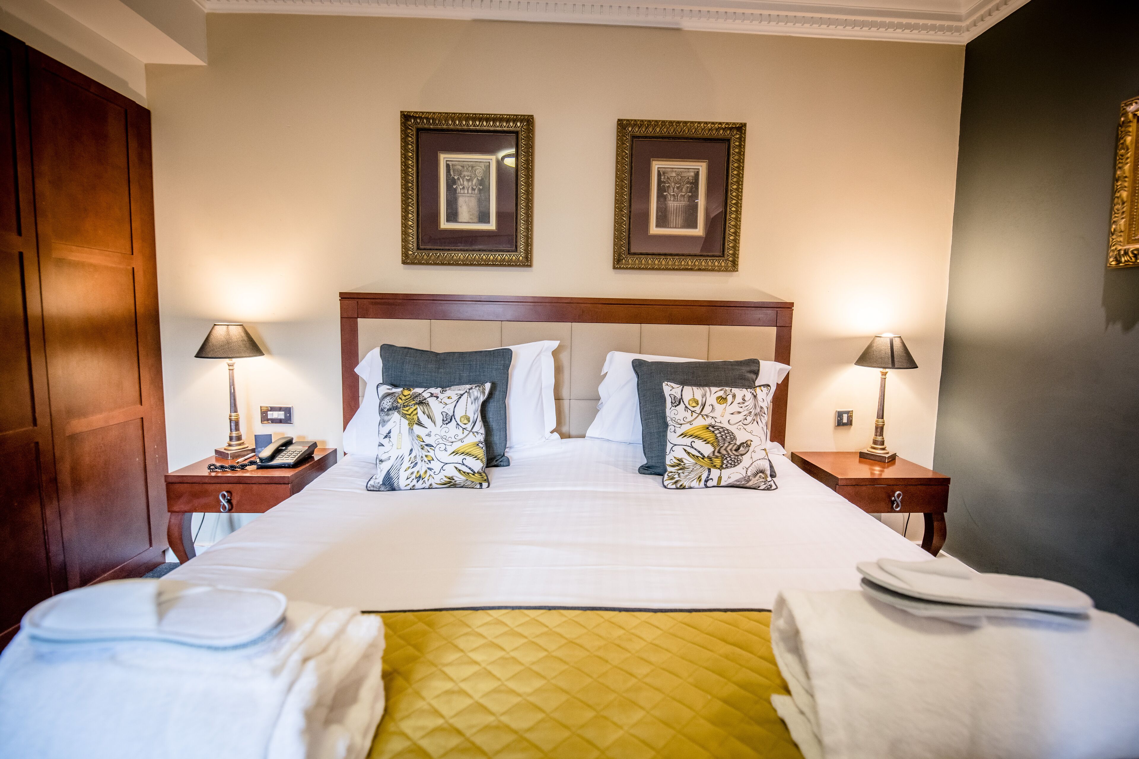 A neatly made bed with a quilted yellow accent, two bedside tables with lamps, and framed pictures on the wall.