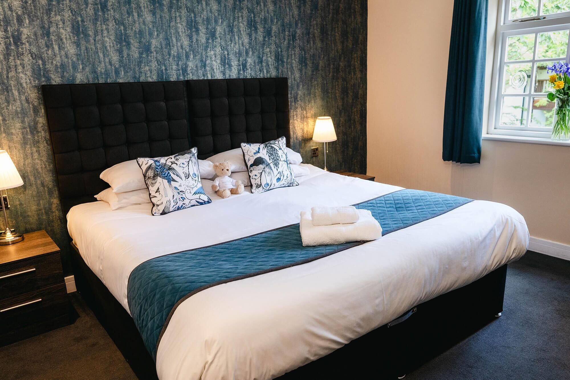 A neatly made bed with a dark headboard, white bedding, and a teal accent. There are two bedside lamps and a window with curtains.