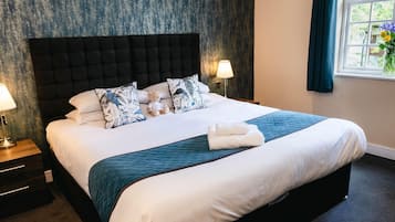 Un lit bien fait, avec une tête de lit sombre, une literie blanche et un coussin ou un élément décoratif de couleur turquoise. On y trouve deux lampes de chevet et une fenêtre avec des rideaux.