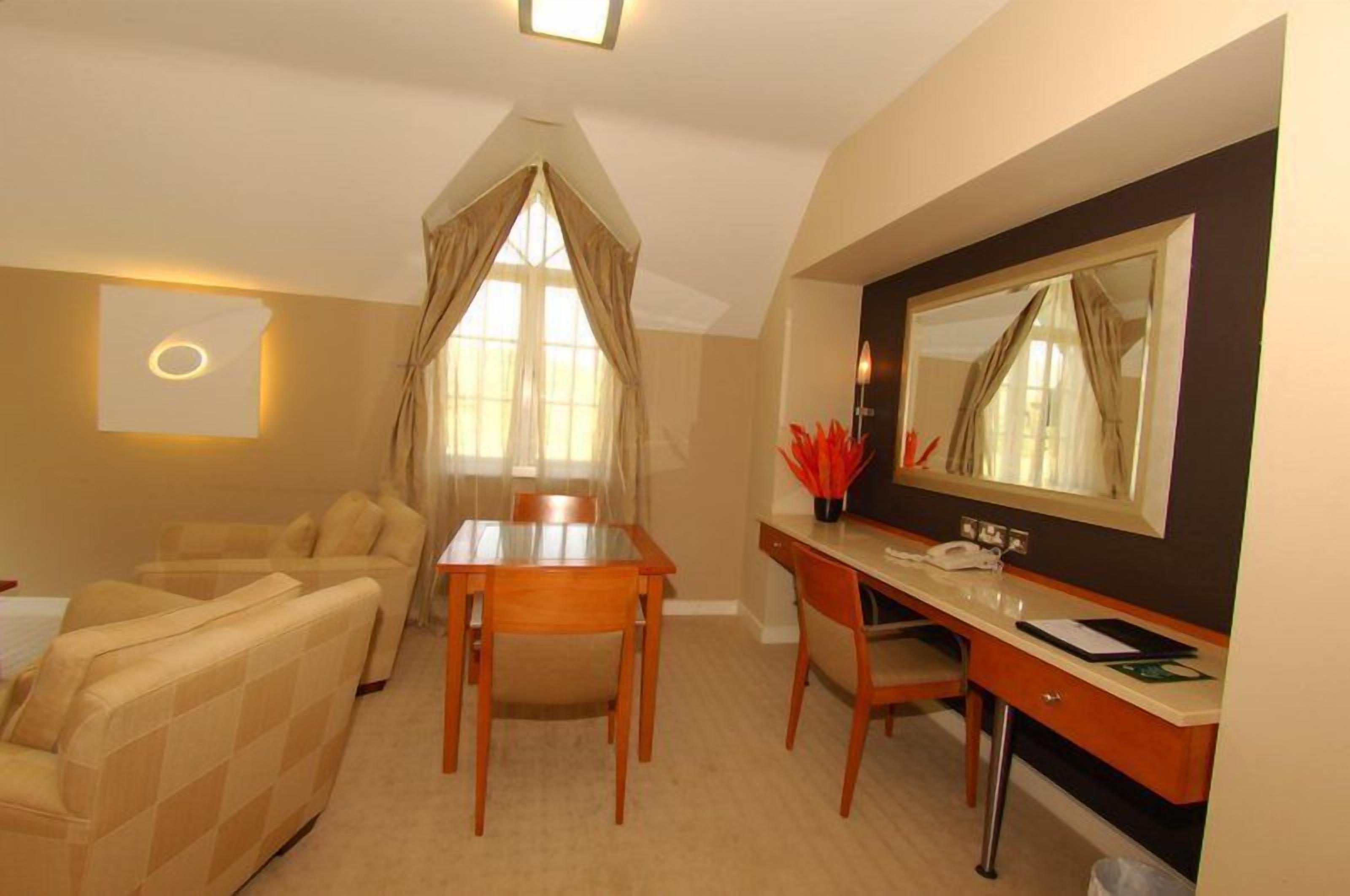 A hotel room with a dining table, a sofa, a desk with a mirror, and a window with curtains.