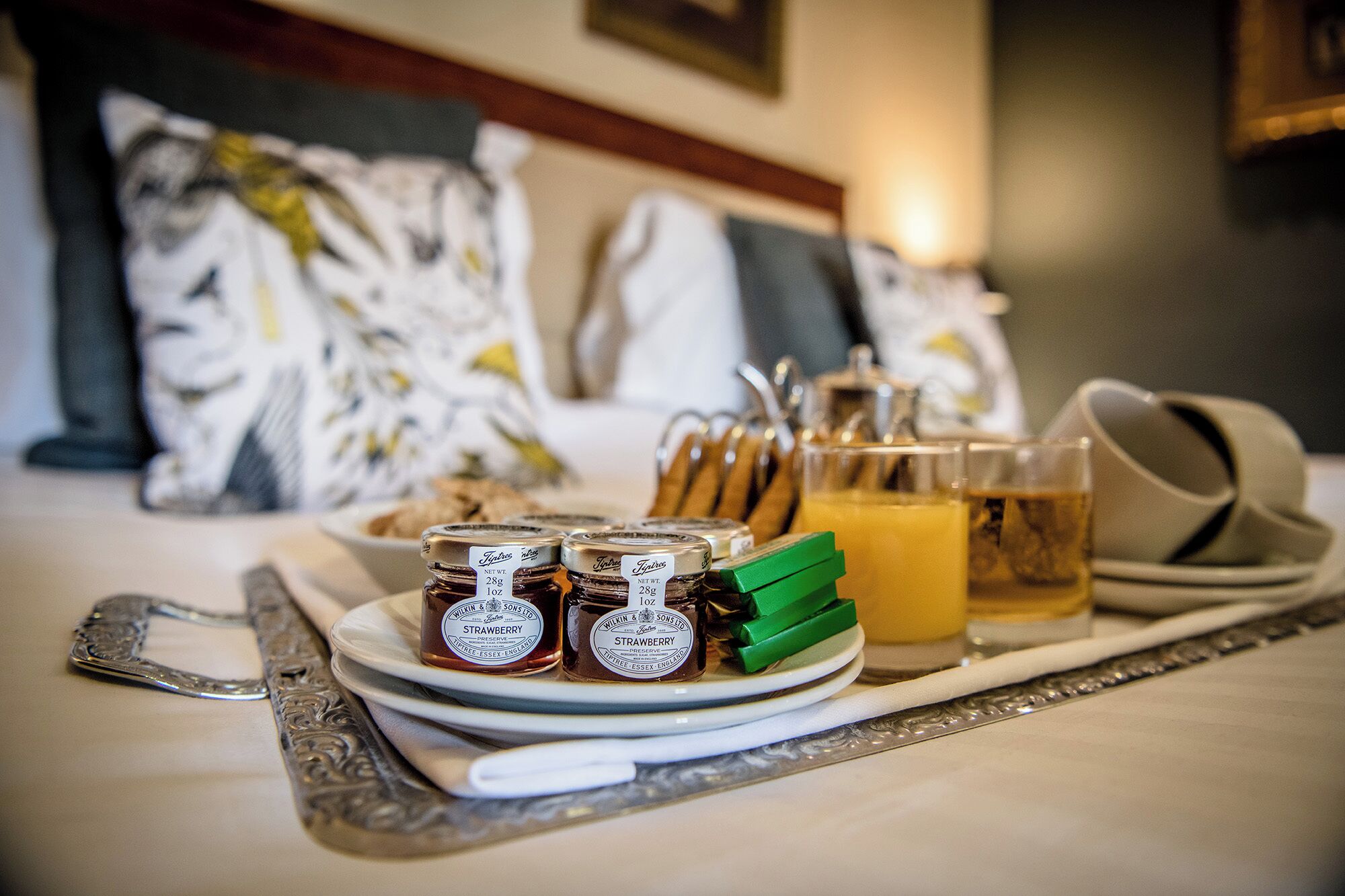 A tray with jam jars, a glass of juice, and a pitcher on a table in a hotel room.