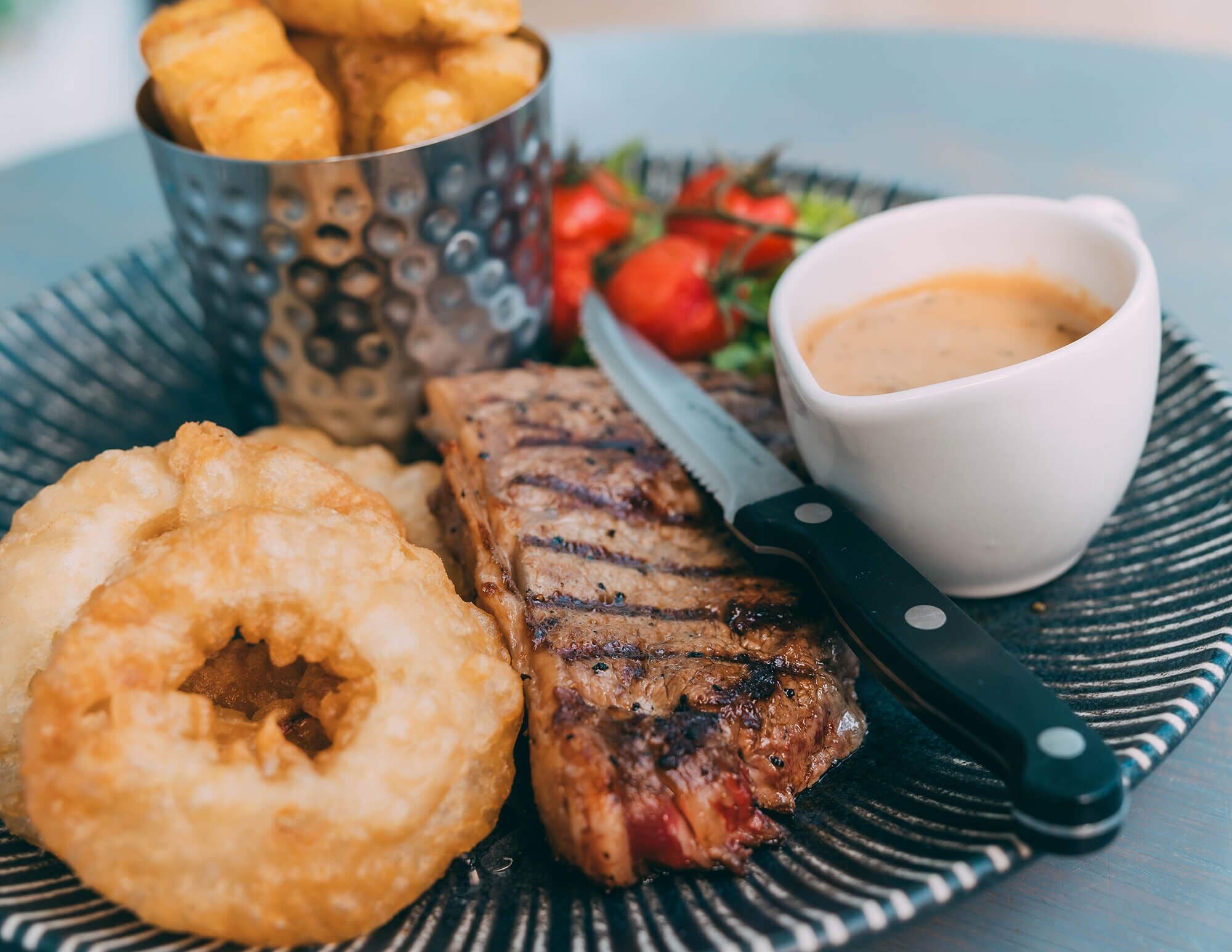 A plate with grilled steak, onion rings, and a cup of sauce.