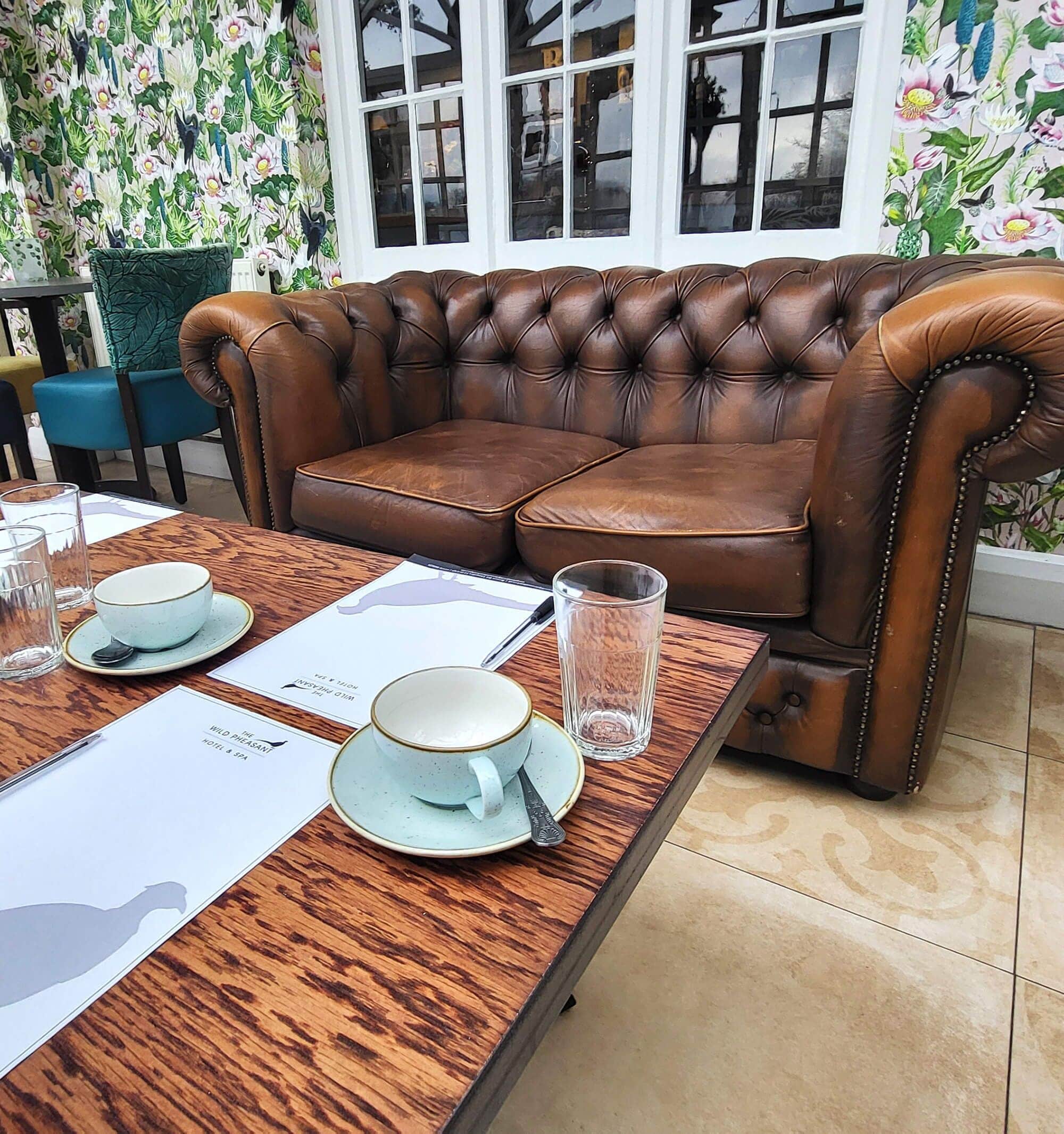 A dining table set with a teapot, cups, and a glass pitcher, with a brown leather sofa and floral wallpaper in the background.