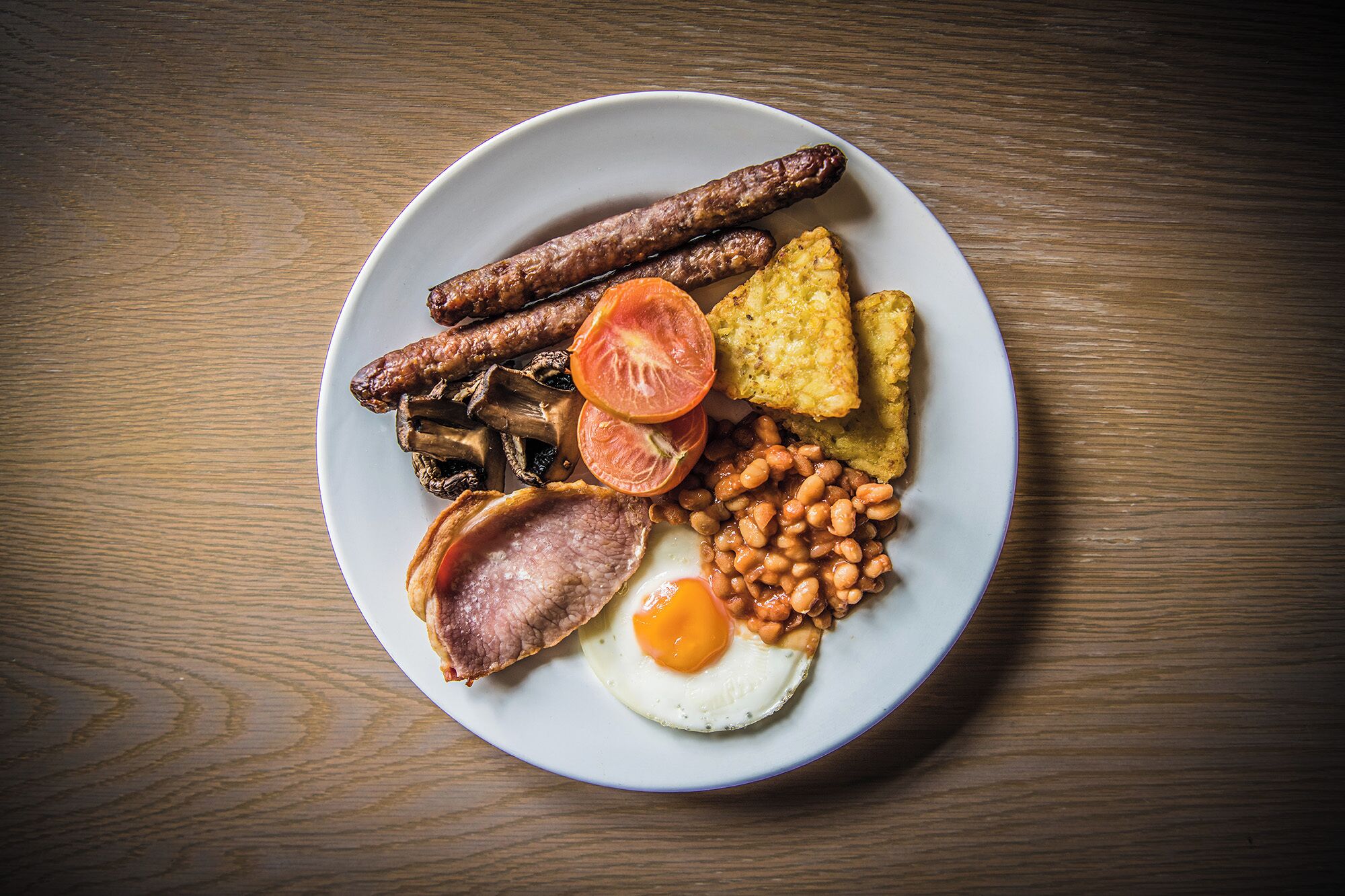 A plate of breakfast food including sausages, mushrooms, tomatoes, beans, fried eggs, and toast.