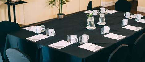 Un salón de conferencias con mantel negro, tazas blancas y cristalería dispuesta para una reunión.