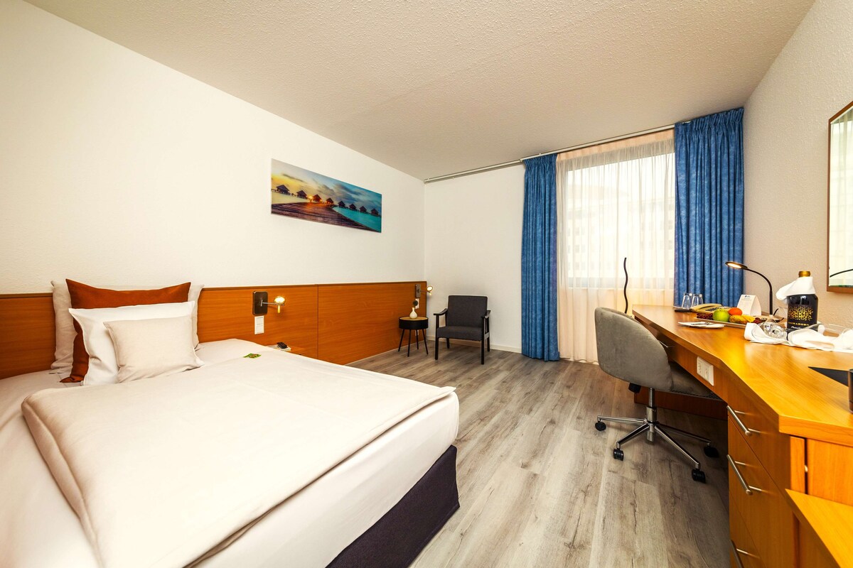 A hotel room with a bed, a desk, a chair, a small table, and a painting on the wall.