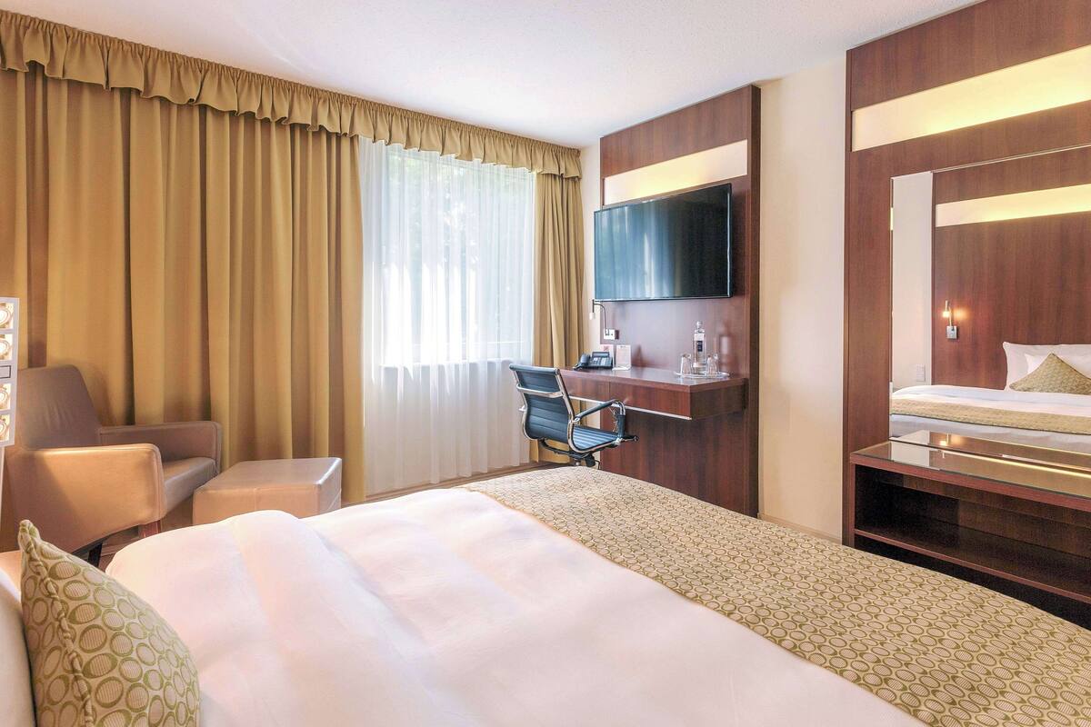 A hotel room with a large bed, a desk with a flat-screen TV, a chair, and a window with curtains.