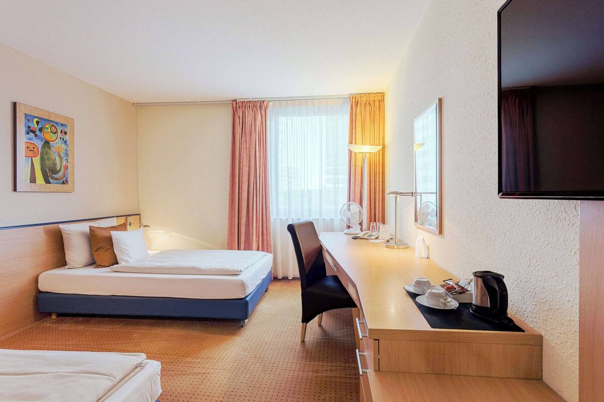 A hotel room with a bed, a desk with a chair, a lamp, a TV, and a painting on the wall.