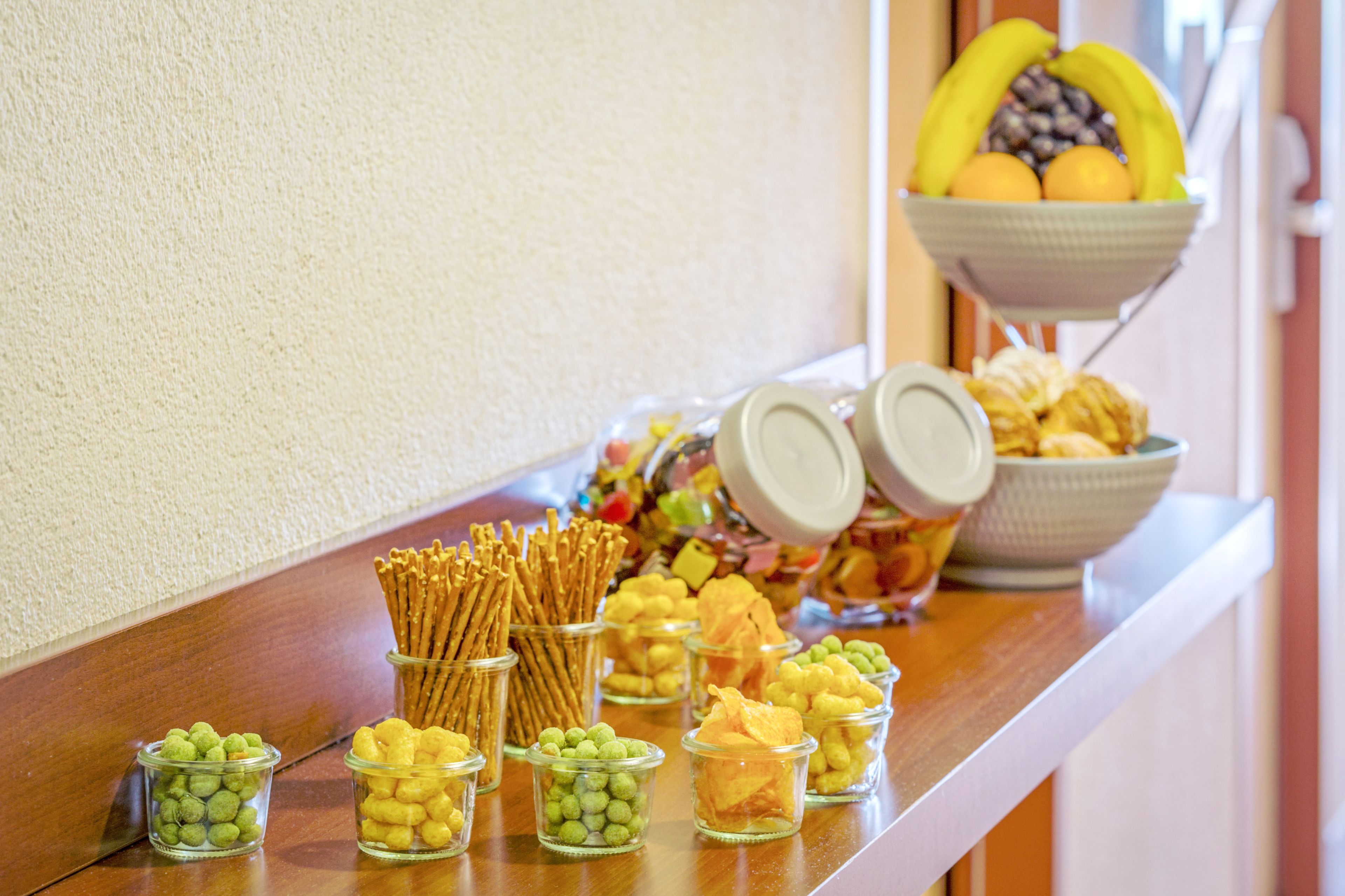 En buffetbod med forskellige spiser, blandt andet kringler, grønne og gule snacks samt en skål med frugt.