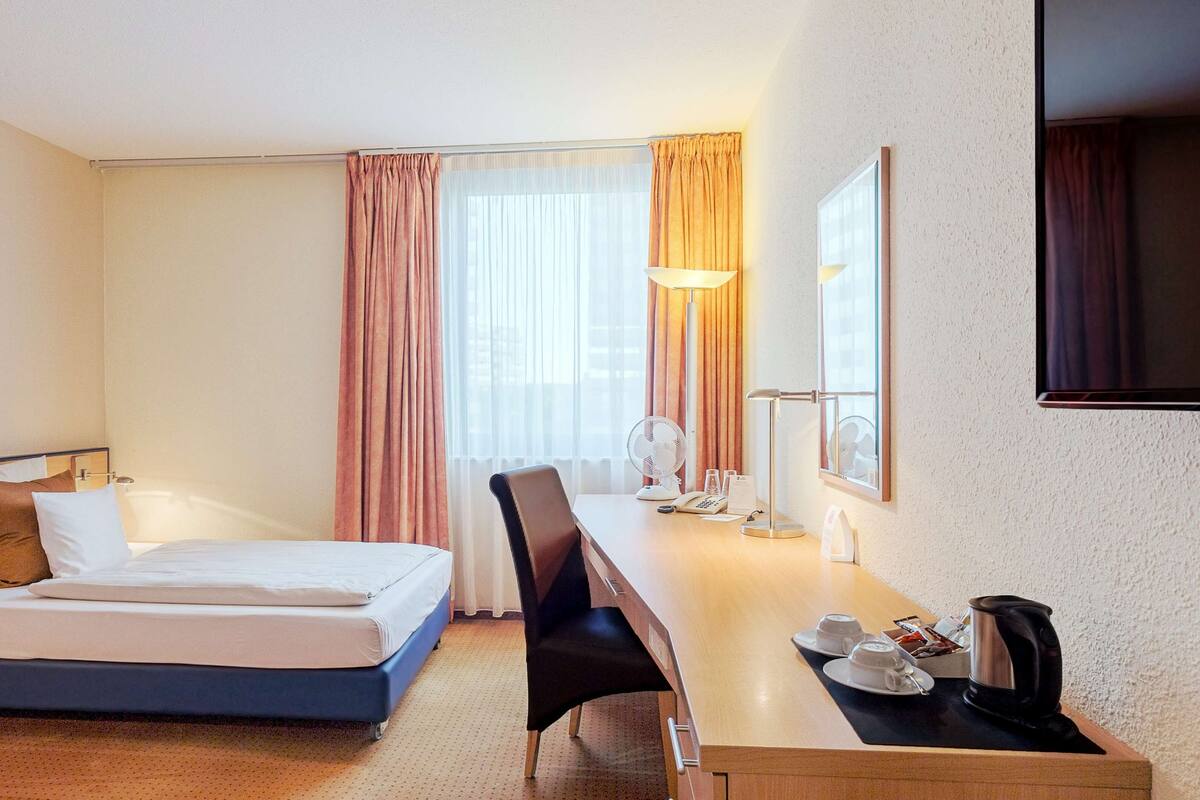 A hotel room with a bed, desk, chair, and a painting on the wall.