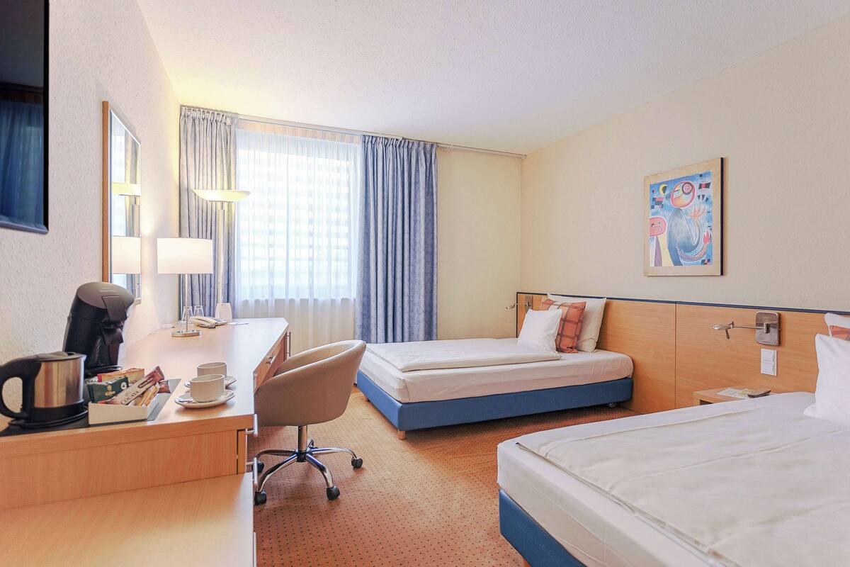 A hotel room with two beds, a desk with a coffee maker, a chair, and a painting on the wall.