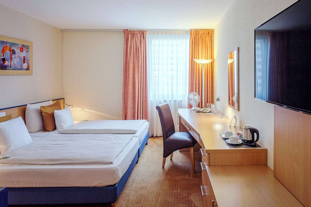A hotel room with two beds, a desk with a chair, a flat-screen TV, and a window with curtains.