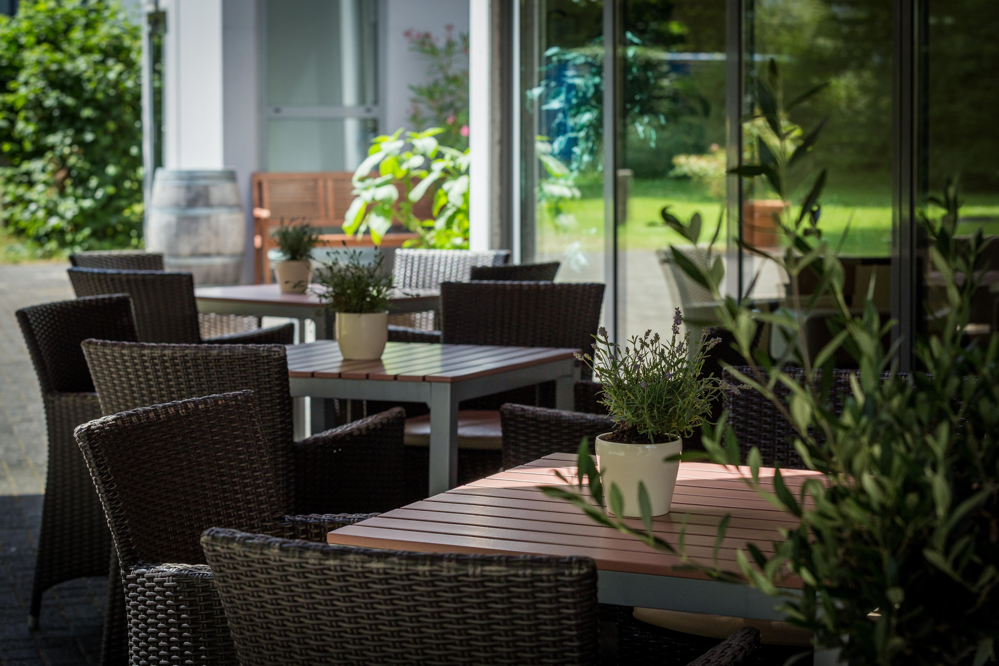 Ein Sitzbereich im Freien mit Korbstühlen und Tischen, Topfpflanzen und Blick auf einen Garten.