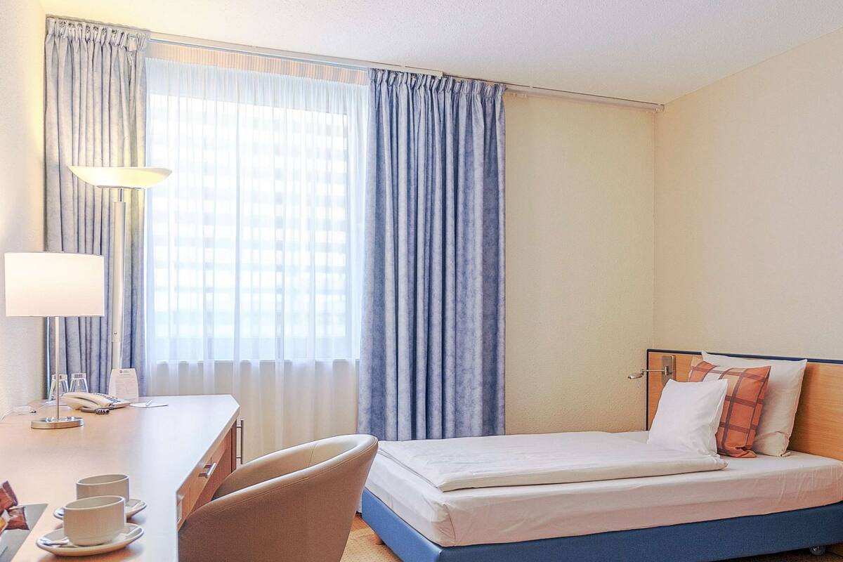 A hotel room with a bed, a desk with a coffee maker, a chair, and a large window with curtains.