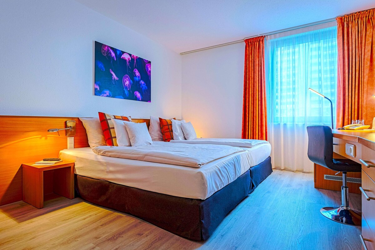 A hotel room with a large bed, a desk with a chair, a nightstand, a painting on the wall, and a window with curtains.