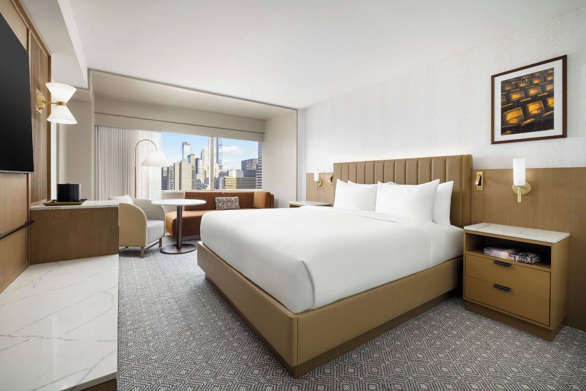 Ein Hotelzimmer mit einem großen Bett, einem Schreibtisch, einem Stuhl, einem kleinen Tisch und einem Fenster mit Blick auf die Stadt.