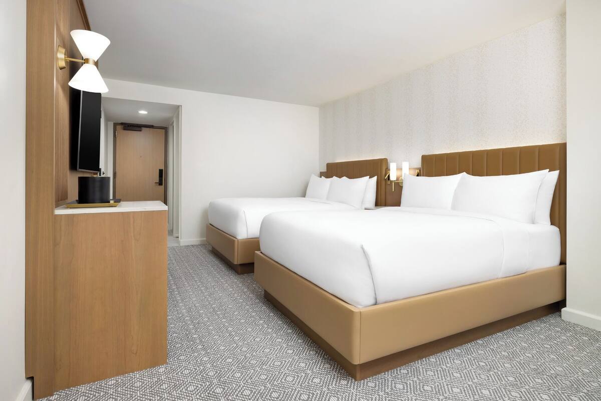Ein Hotelzimmer mit zwei Betten, einem Holztisch, einem Flachbildfernseher und einem gemusterten Teppich.
