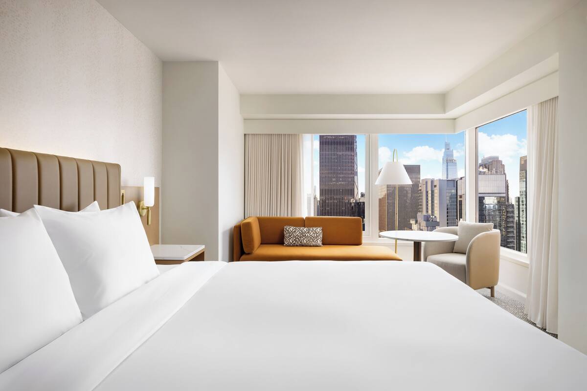 Ein Hotelzimmer mit einem großen Bett, einer Couch, einem kleinen Tisch und einem Sessel. Ein großes Fenster bietet Blick auf die Stadt.