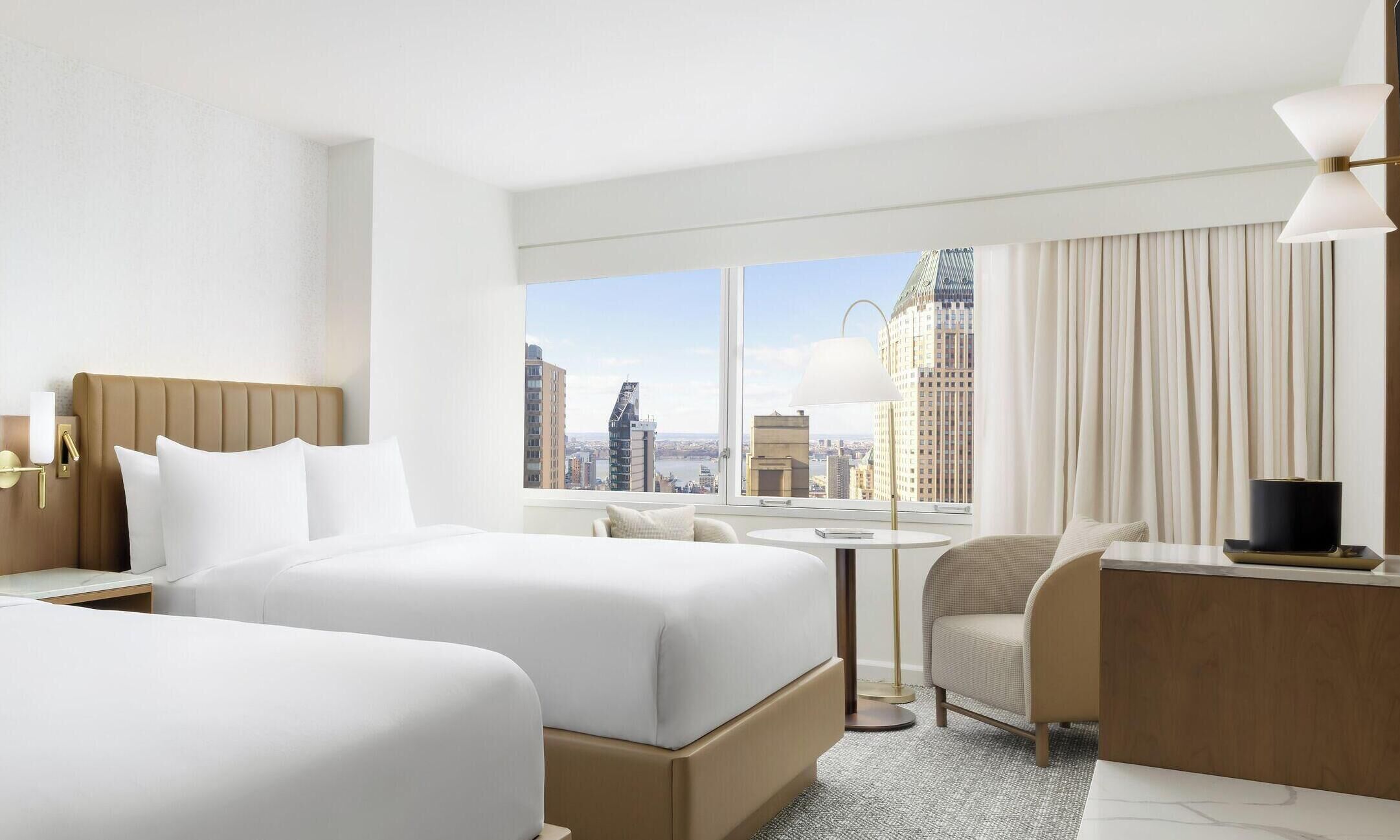 A hotel room with two beds, a desk, a chair, a small table, and a view of the cityscape.