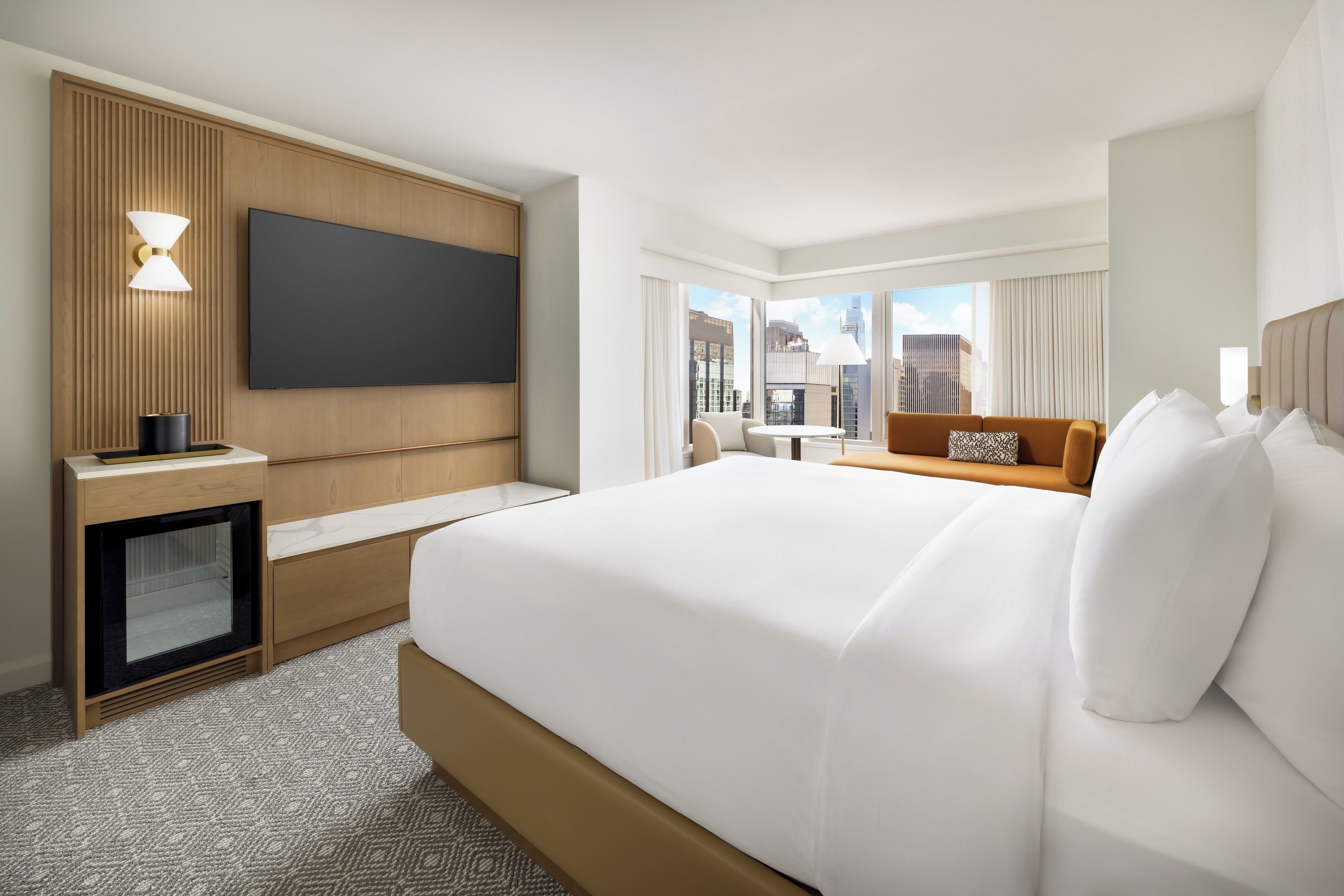 Ein Hotelzimmer mit einem großen Bett, einem an der Wand befestigten Fernseher, einem Schreibtisch mit Lampe und Blick auf die Stadt durch große Fenster.