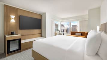 Ein Hotelzimmer mit einem großen Bett, einem an der Wand befestigten Fernseher, einem Schreibtisch mit Lampe und Blick auf die Stadt durch große Fenster.