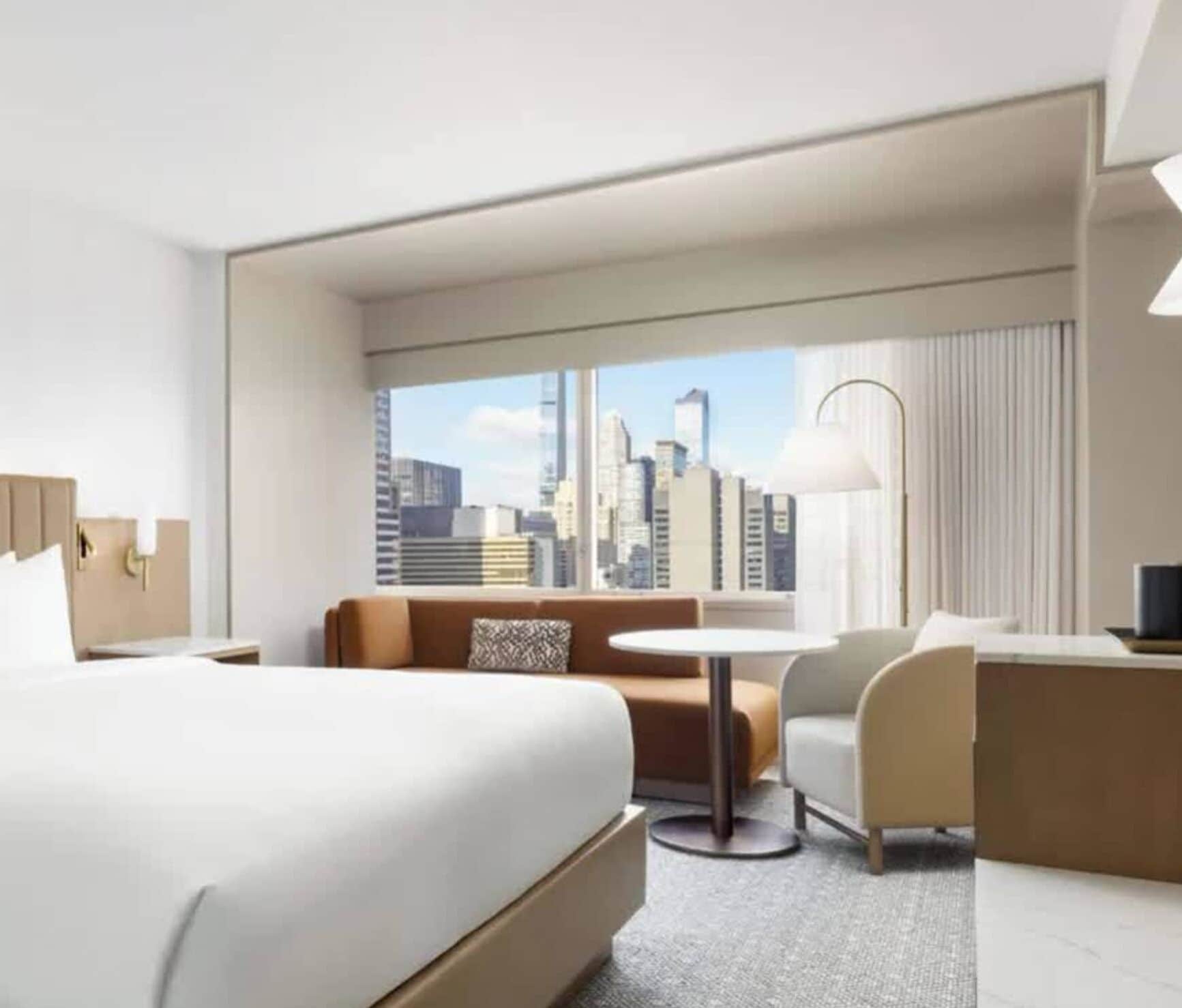 Habitación de hotel con una cama grande, un sofá, una pequeña mesa redonda y una silla. Cuenta con un amplio ventanal con vista a la ciudad.