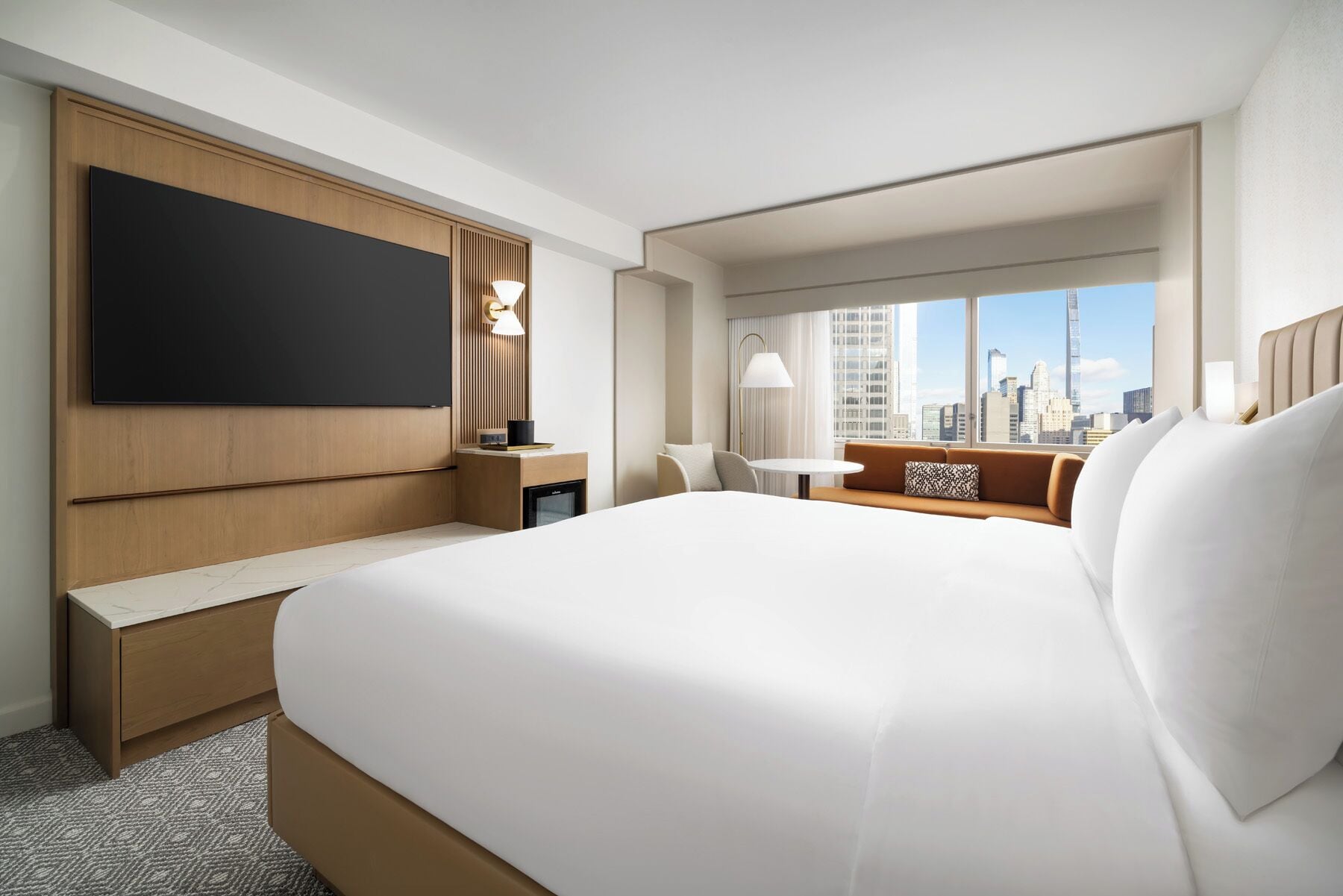 Ett hotellrum med en stor säng, en platt-TV, ett skrivbord, en stol, ett litet bord och utsikt över stadens silhuett.