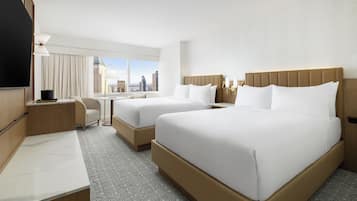 A hotel room with two beds, a desk, a chair, and a large window offering a city view.