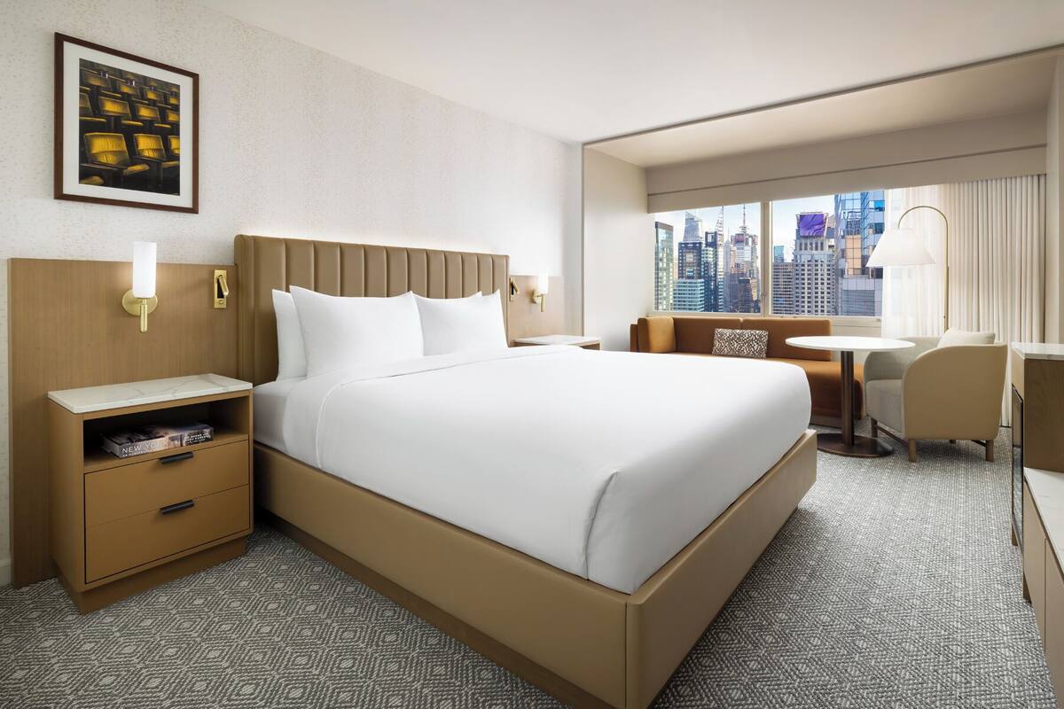 Ein Hotelzimmer mit einem großen Bett, einem Nachttisch mit Lampe, einem kleinen Schreibtisch mit Stuhl und Blick auf die Stadt durch das Fenster.
