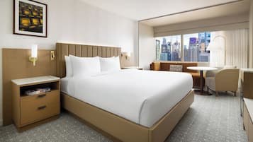 Een hotelkamer met een groot bed, een nachtkastje met een lamp, een klein bureau met een stoel, en uitzicht op de stad door het raam.