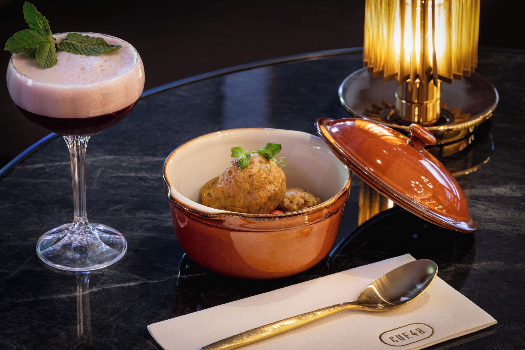 A dessert setting with a glass of a pinkish drink, a bowl of food with a lid, and a spoon on a napkin.