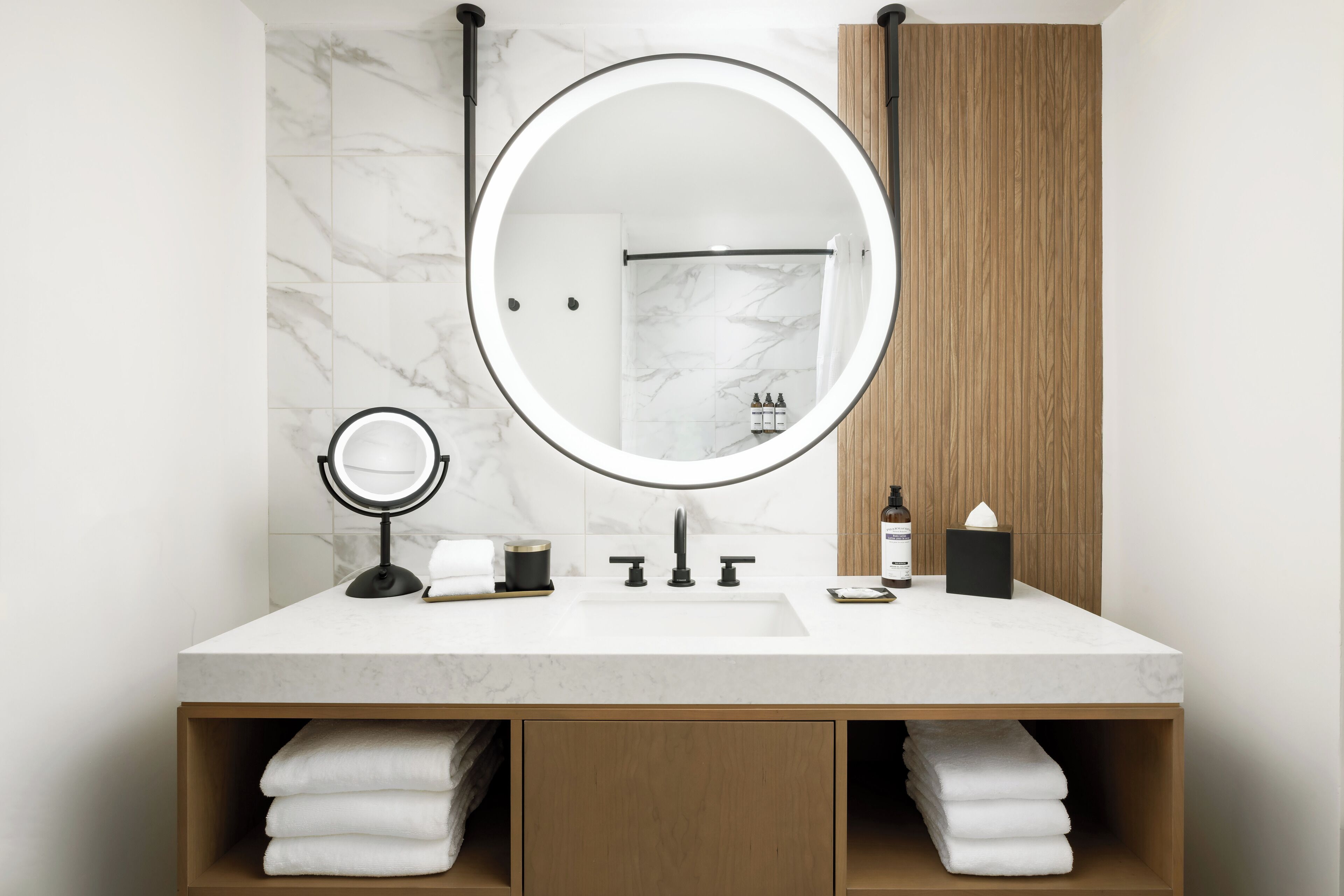 A bathroom with a marble wall, a round mirror with lights, a sink, and wooden cabinets.