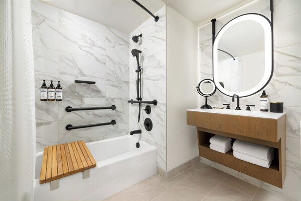 A modern bathroom with a marble shower, a wooden bench, a vanity with a mirror, and toiletries.