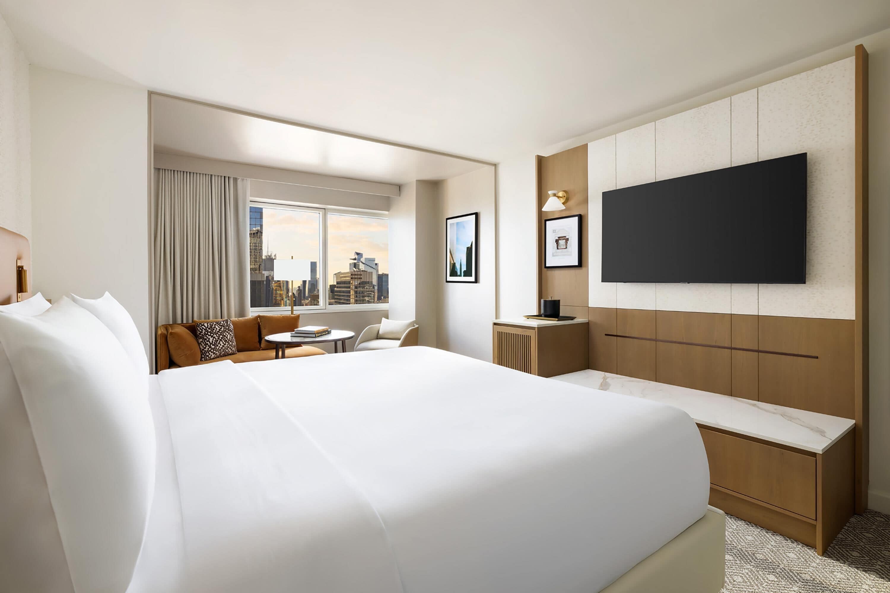 A hotel room with a large bed, a seating area with a sofa and chairs, a television mounted on the wall, and a window with a city view.