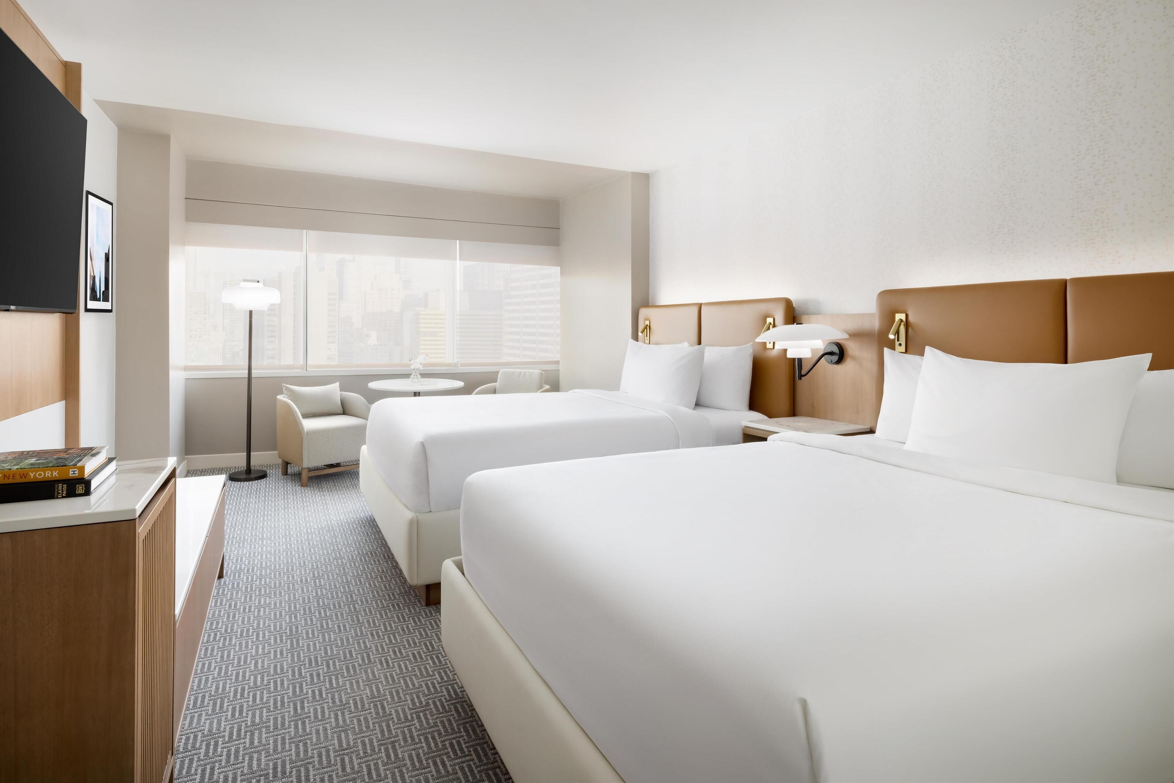 A hotel room with two beds, a desk, a chair, a lamp, and a window with blinds.