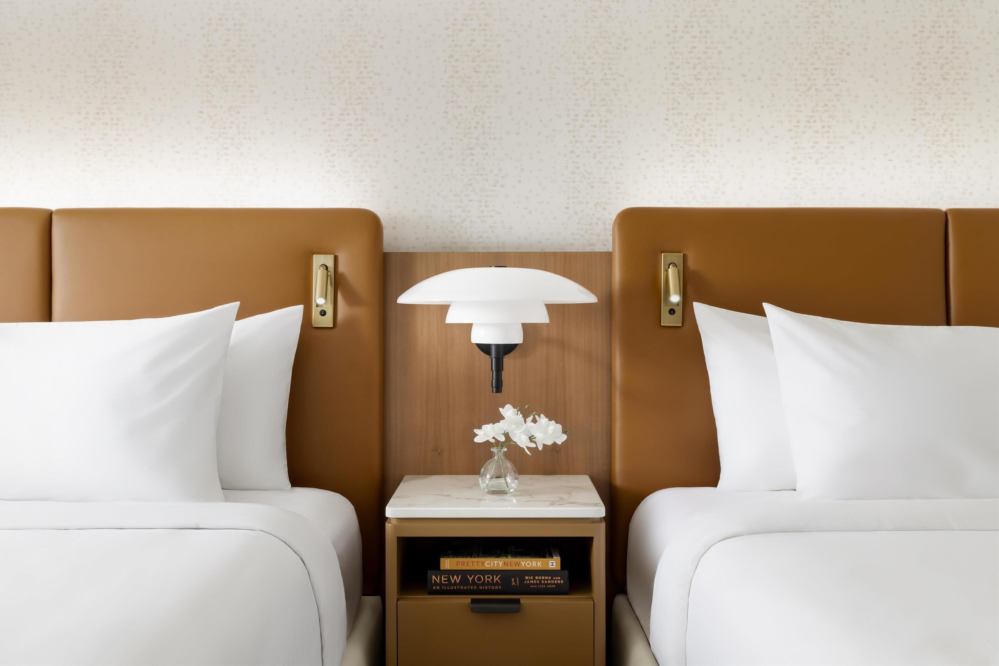 A hotel room with two beds, a nightstand with a lamp, a vase with flowers, and books.